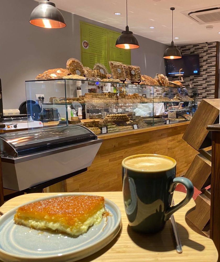 A slice of syrup-soaked semolina cake beside a latte in a green mug, the pastry counter and bread shelves behind.
